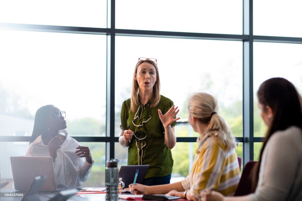 Four women in a bright meeting room. One woman stands, speaking and gesturing, while three others sit at a table, listening attentively. Laptops and notes are on the table.