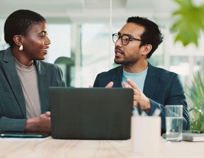 Two professionals, a man and a woman, are seated at a desk, looking at each other and talking. A laptop is between them.