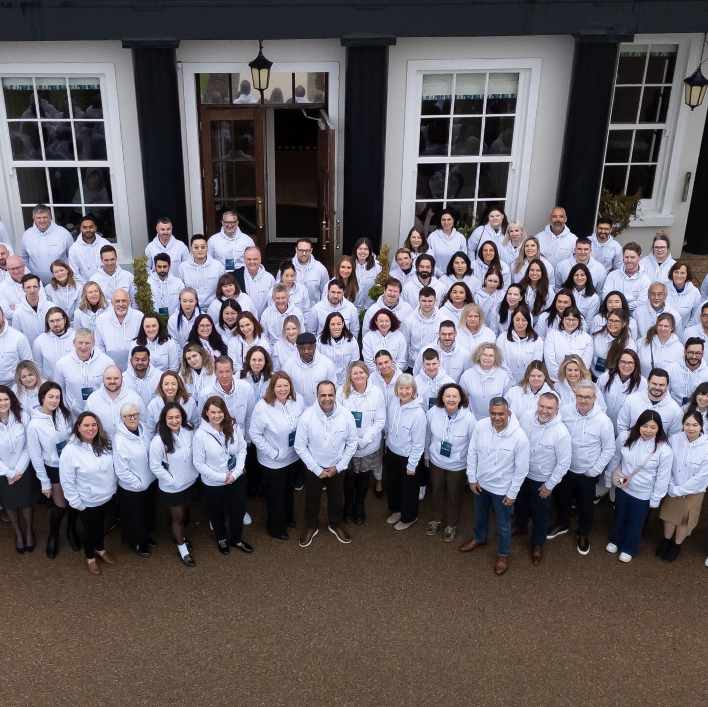 A large group of diverse people, all wearing matching white hooded sweatshirts with a logo, smiling and posing for a group photo outdoors in front of a white building with dark architectural details.
