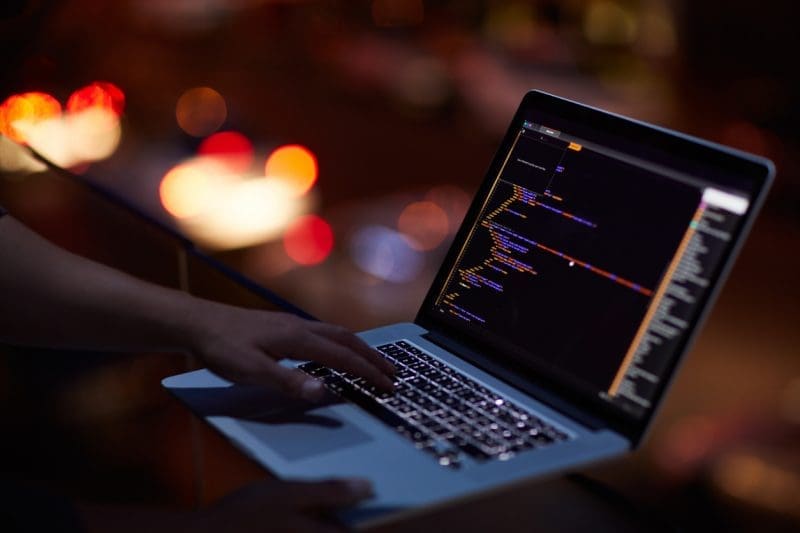 A person's hands type code on a laptop in a dark setting, with blurred red and orange city lights in the background.