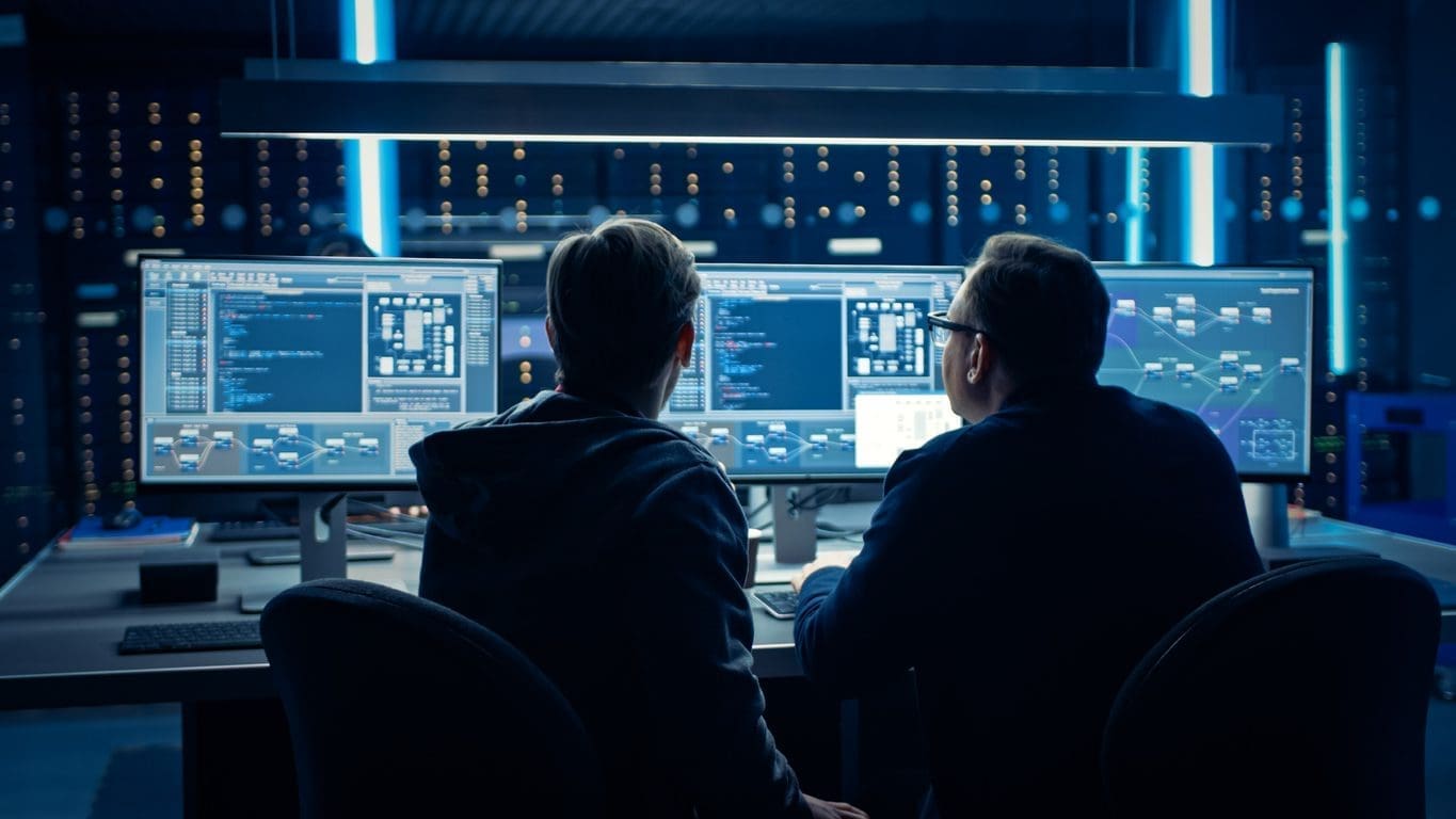 Two men from behind, working on multiple computer screens displaying code and network diagrams in a dark, high-tech server room with blue lights.