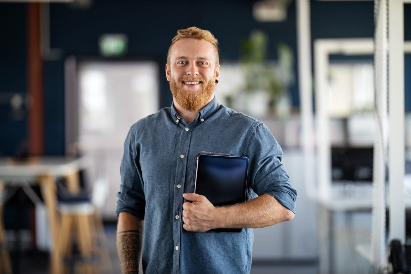 A smiling man with a red beard and a blue shirt holds a tablet in a modern office.