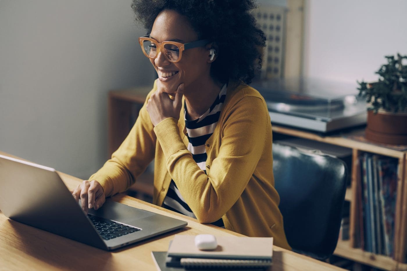 A smiling Black woman with curly hair and orange glasses works on a laptop at a wooden desk, wearing a yellow cardigan.