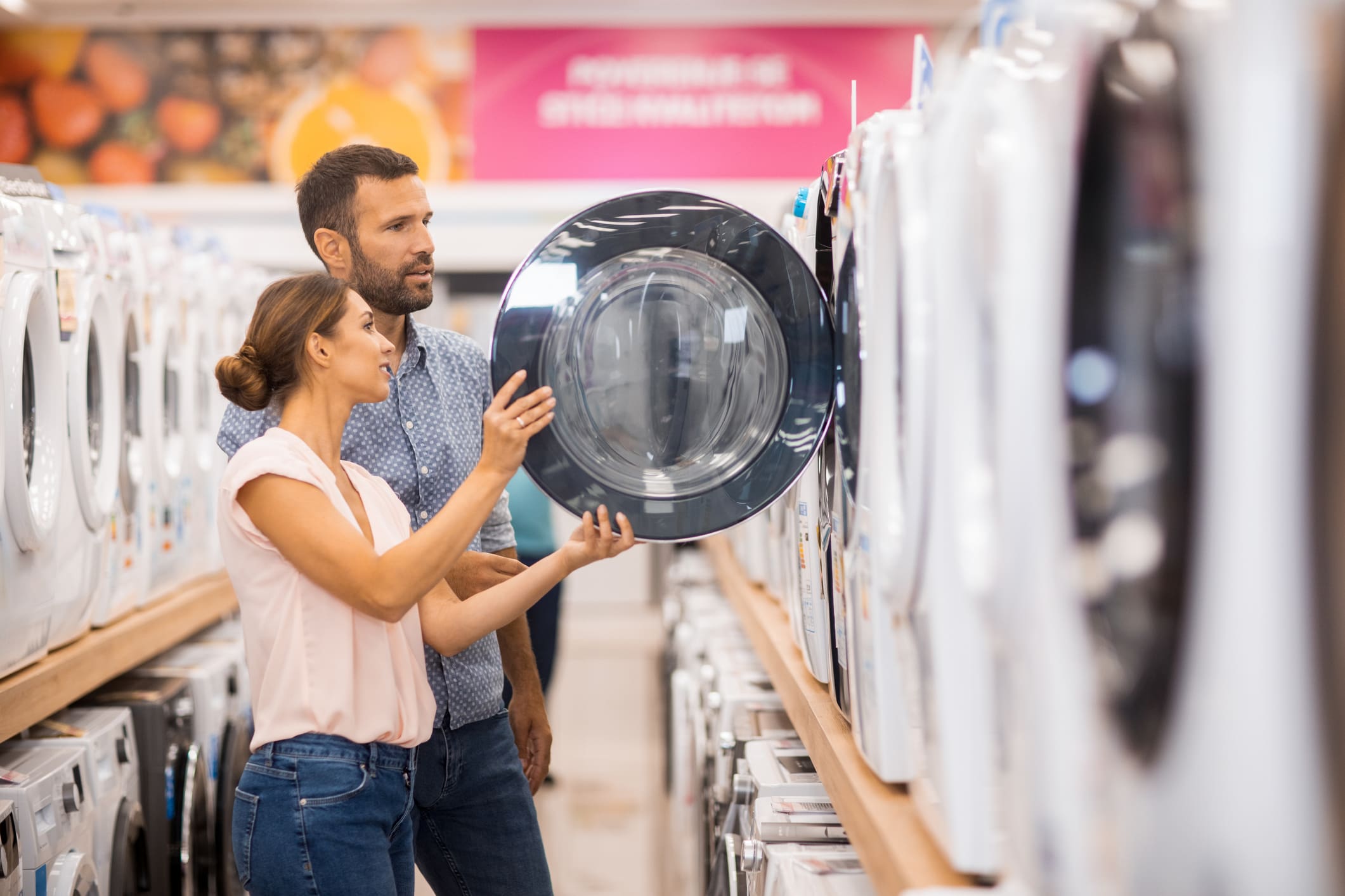 A man and woman examine a washing machine in an appliance store. The woman holds open the dark door of a front-loader while the man looks on.