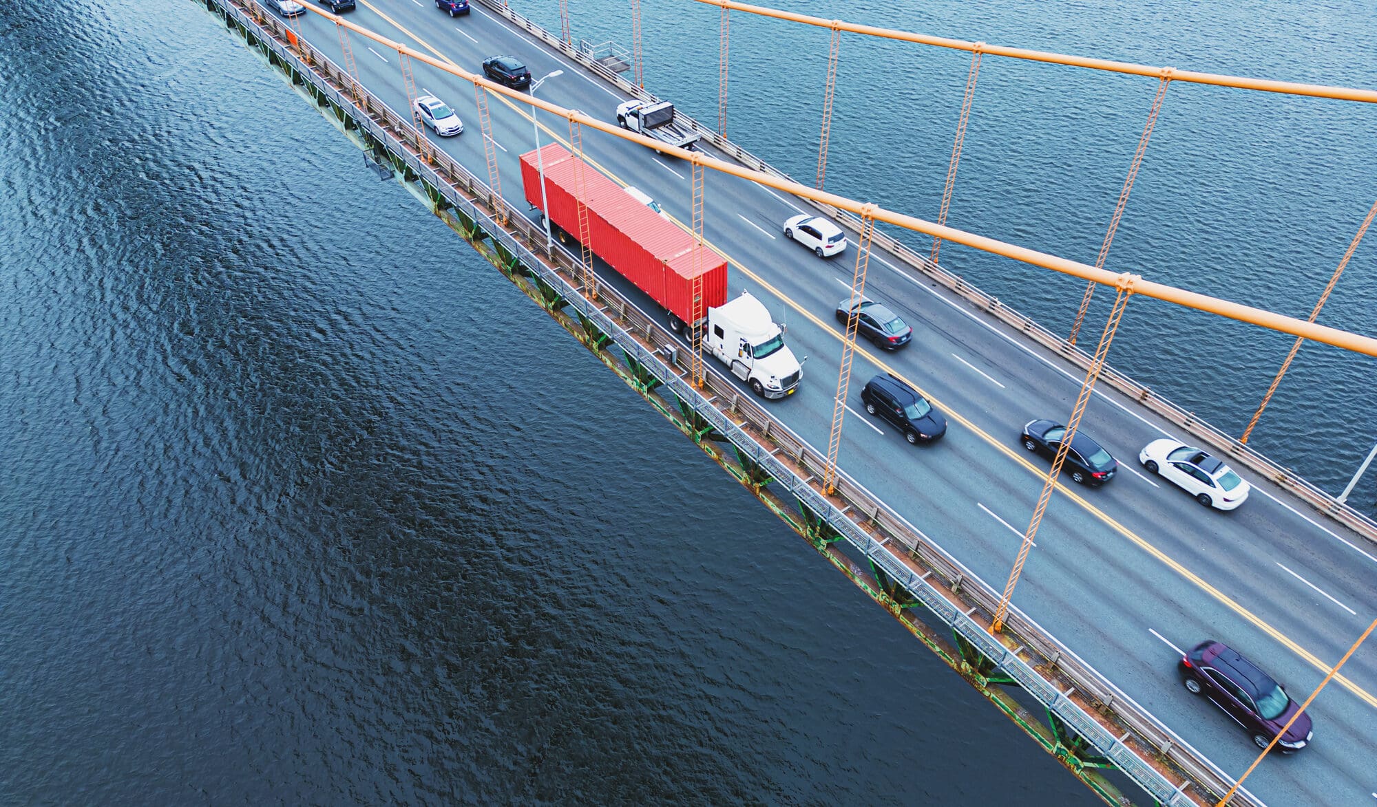 High-angle view of a suspension bridge over dark water. Cars and a large red semi-truck are driving on the bridge, supported by orange cables.