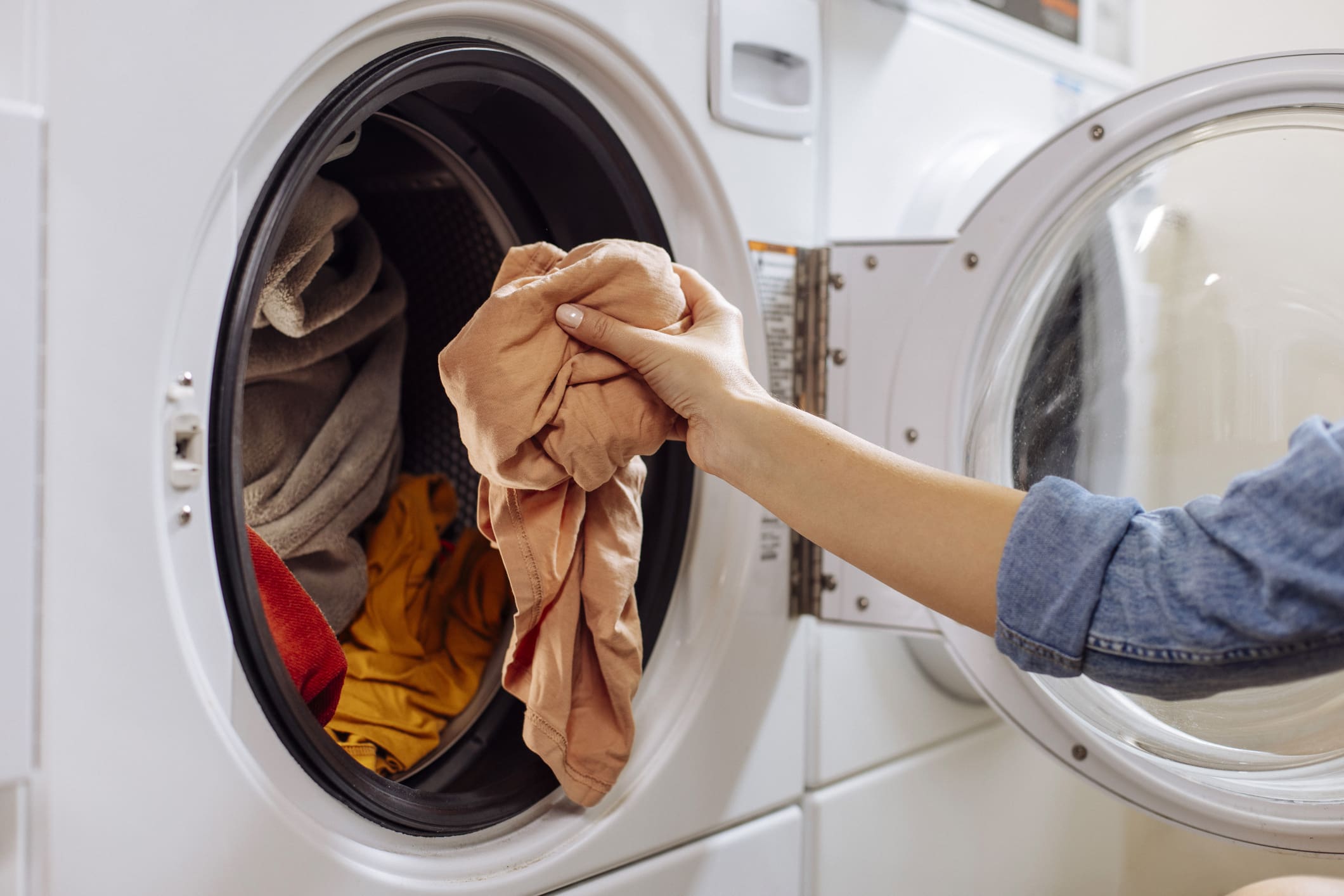 A hand puts a peach-colored garment into an open front-loading washing machine filled with other clothes.