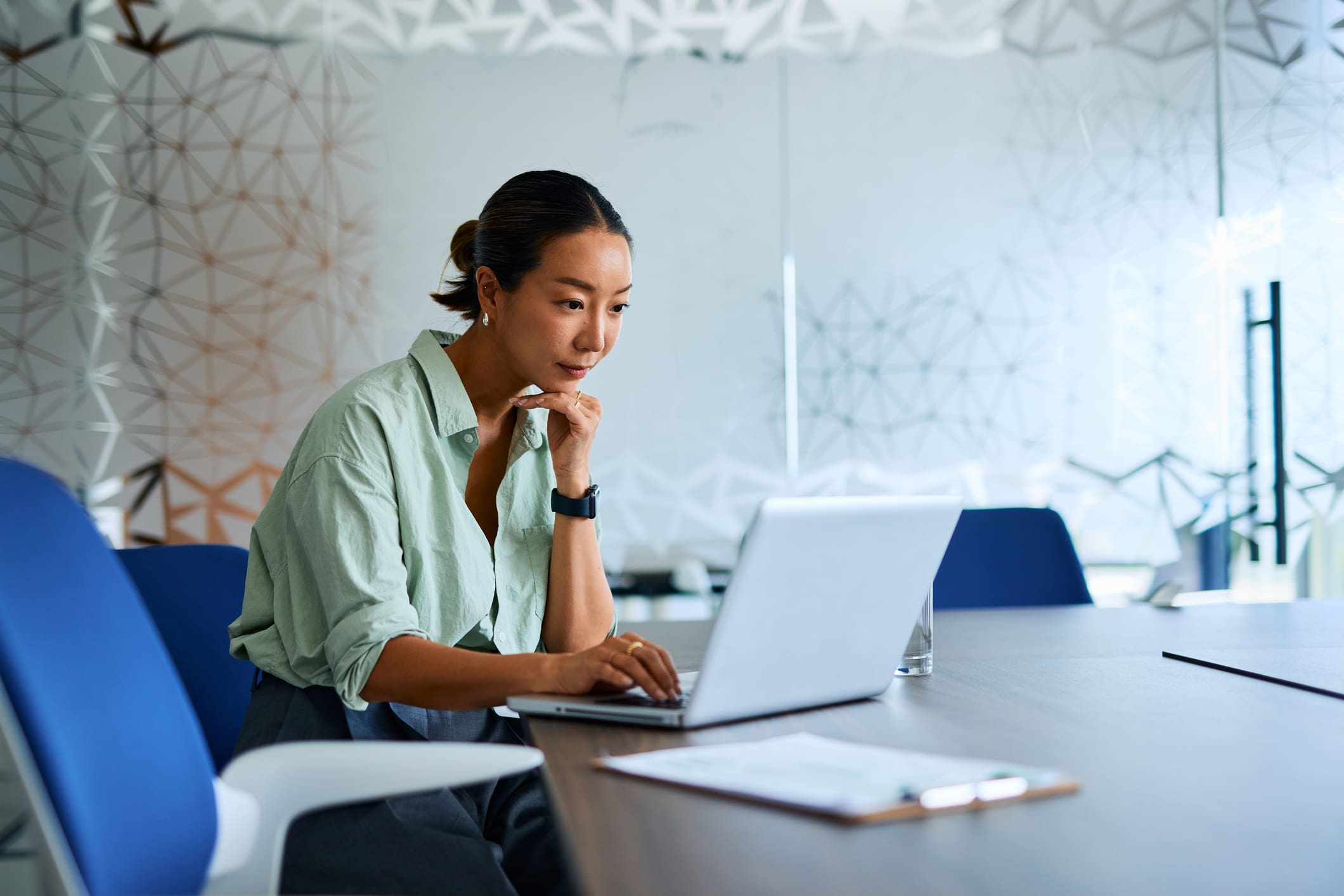 A woman in a green shirt sits at a conference table, typing on a laptop with her hand on her chin, in a modern office with a geometric glass wall.
