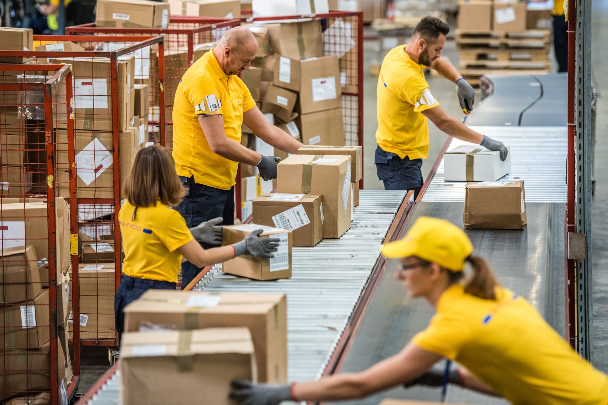 Four logistics workers in yellow uniforms and gloves sort cardboard packages on a conveyor belt in a busy warehouse.