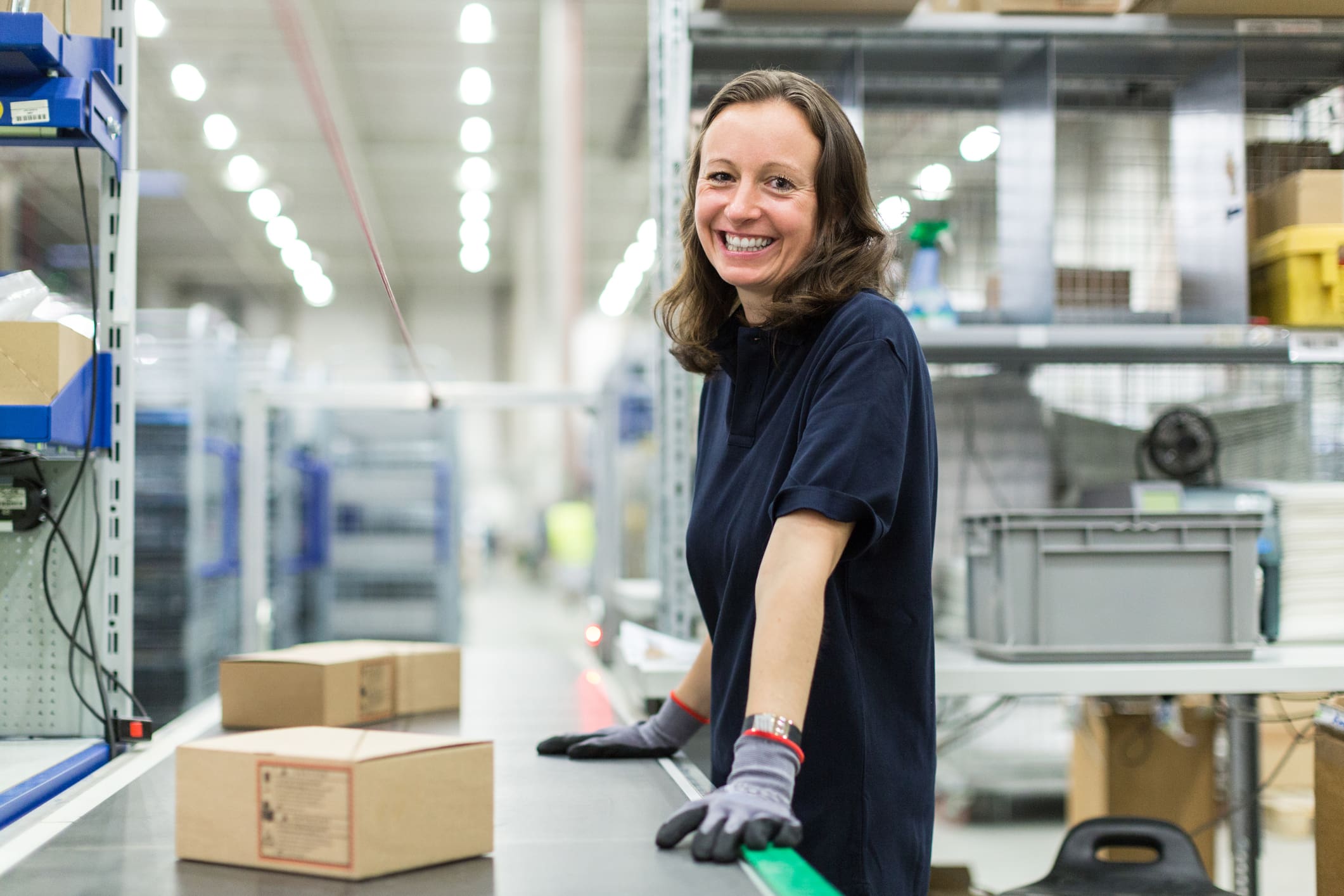 A smiling woman in a dark blue shirt and work gloves stands next to a conveyor belt with boxes in a bright, modern warehouse.
