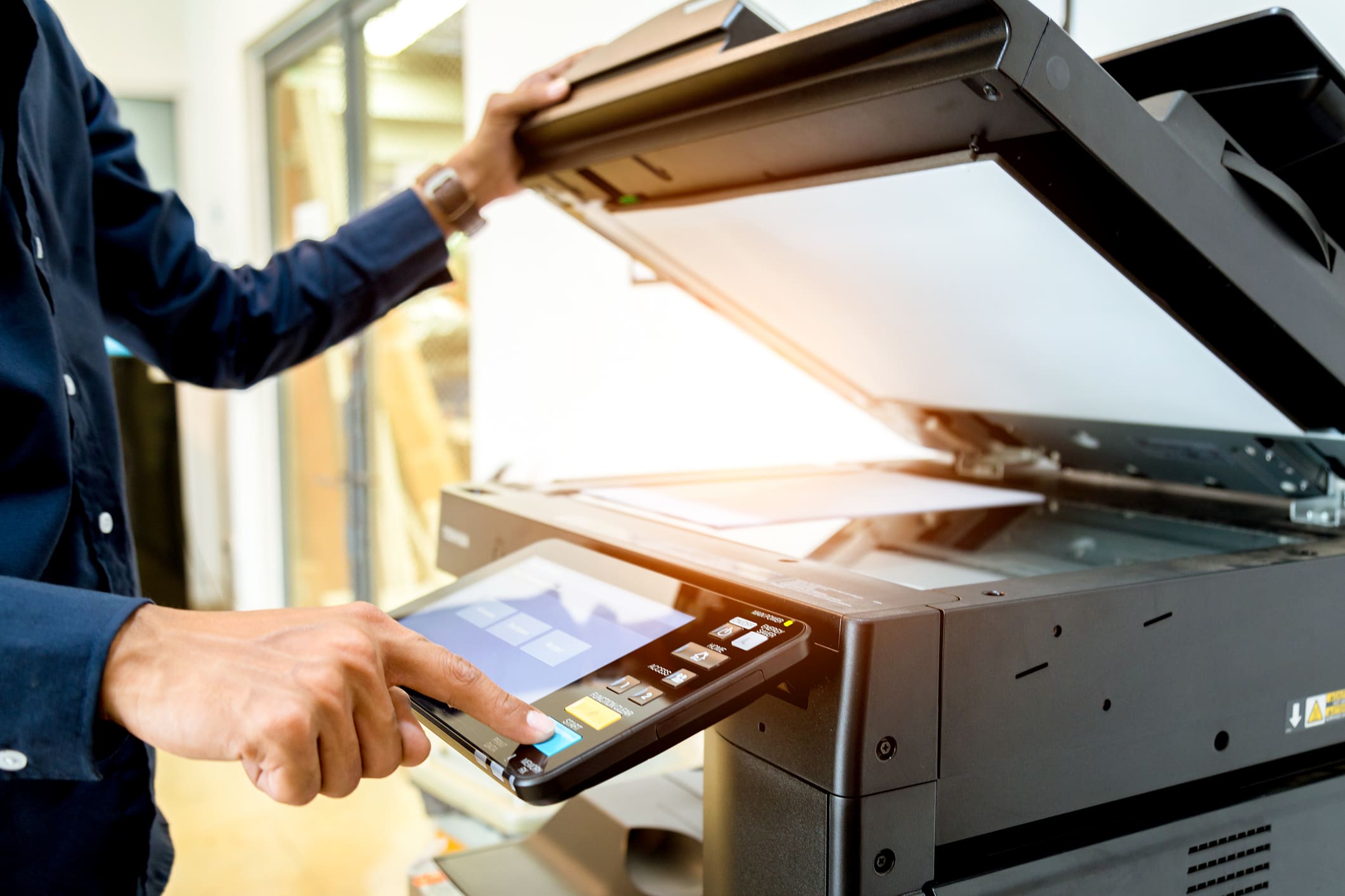 A person in a blue shirt operating a black multifunction printer. One hand lifts the lid while the other presses the 'Start' button on the control panel.