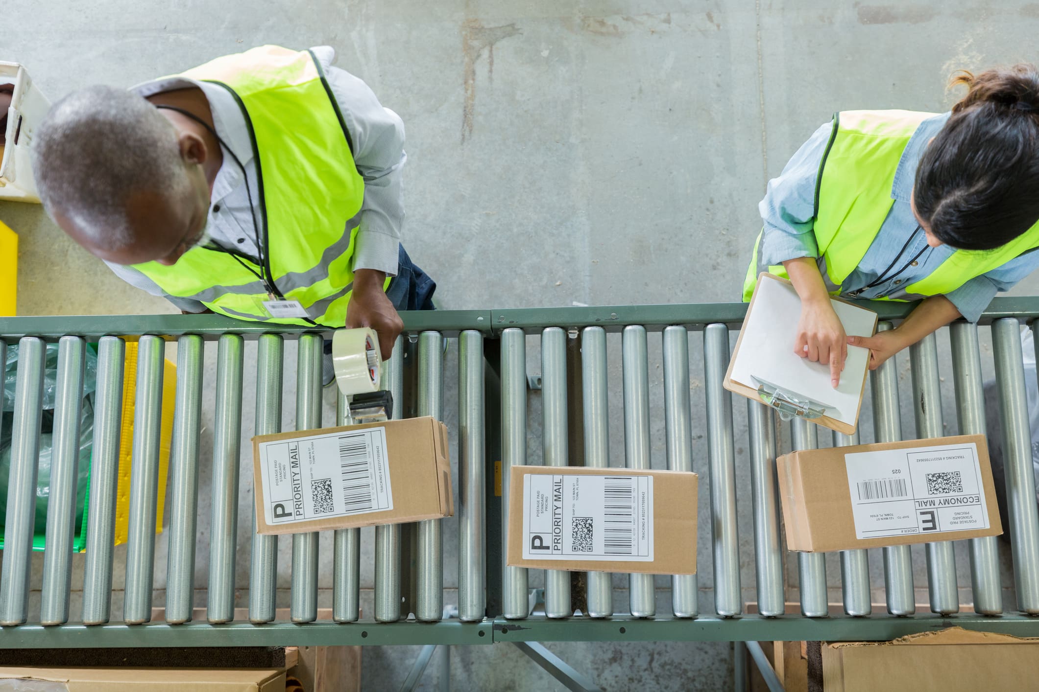 Overhead view of two workers in high-visibility vests processing packages on a conveyor belt in a warehouse. A man holds tape, and a woman holds a clipboard, with "PRIORITY MAIL" and "ECONOMY MAIL" boxes visible.