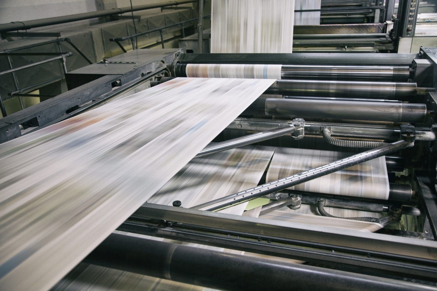 A close-up of an industrial printing press with blurred paper rapidly moving through large metal rollers, conveying high-speed production.