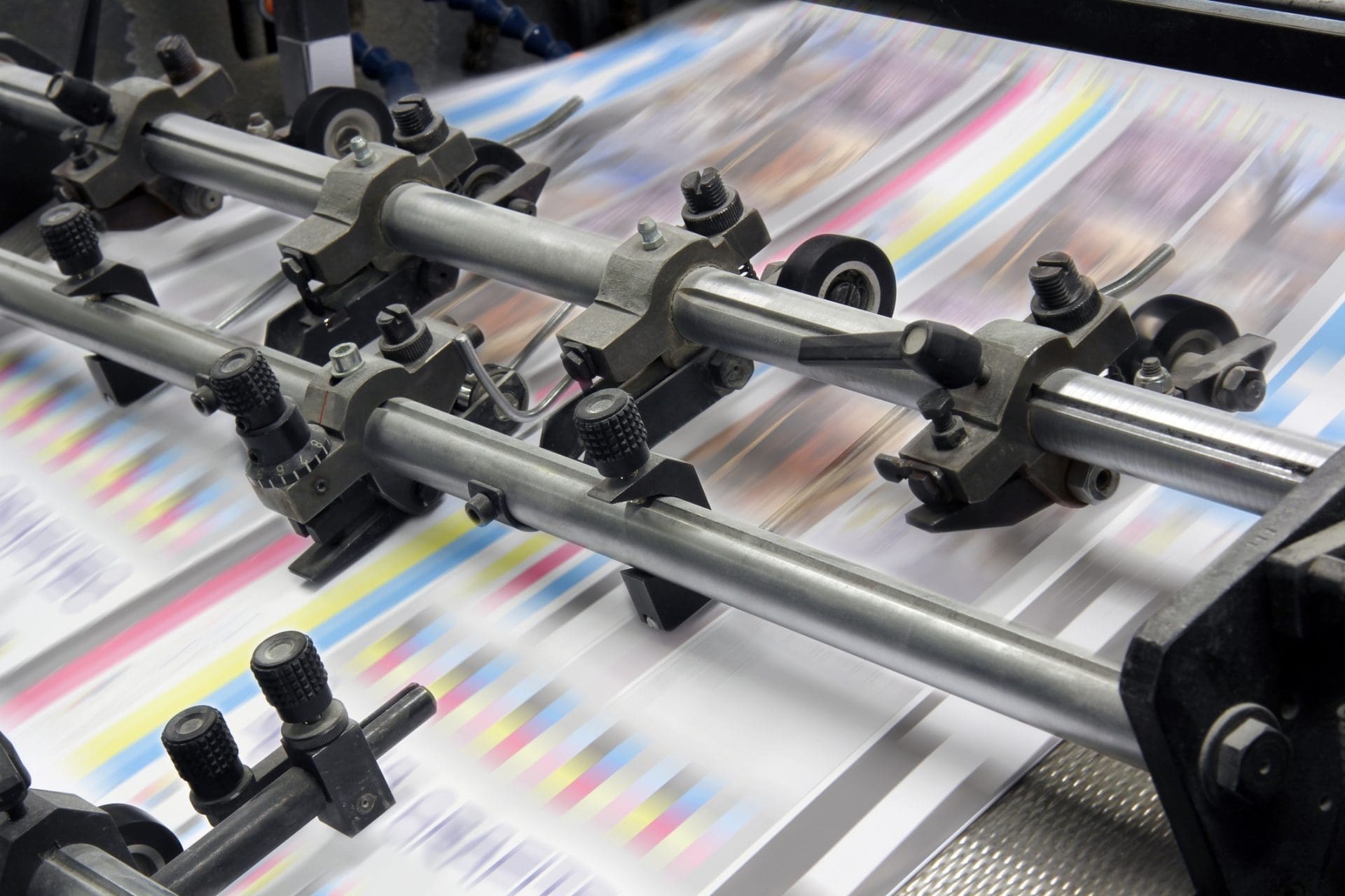 Close-up of an industrial printing press with paper moving rapidly through metal rollers. The paper shows blurred streaks of cyan, magenta, yellow, and black colors, indicating active printing.