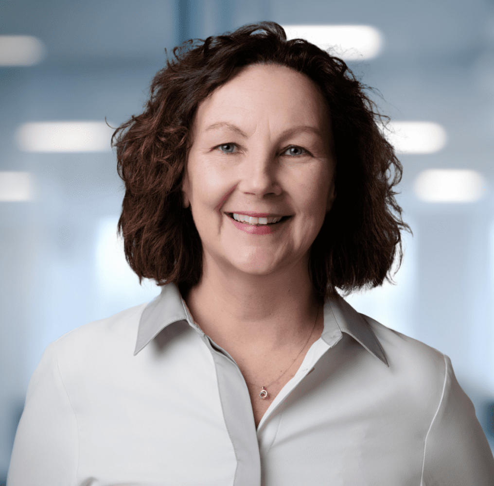 Headshot of a smiling woman with curly brown hair and blue eyes, wearing a light shirt and a necklace, against a blurred office background with a plant.