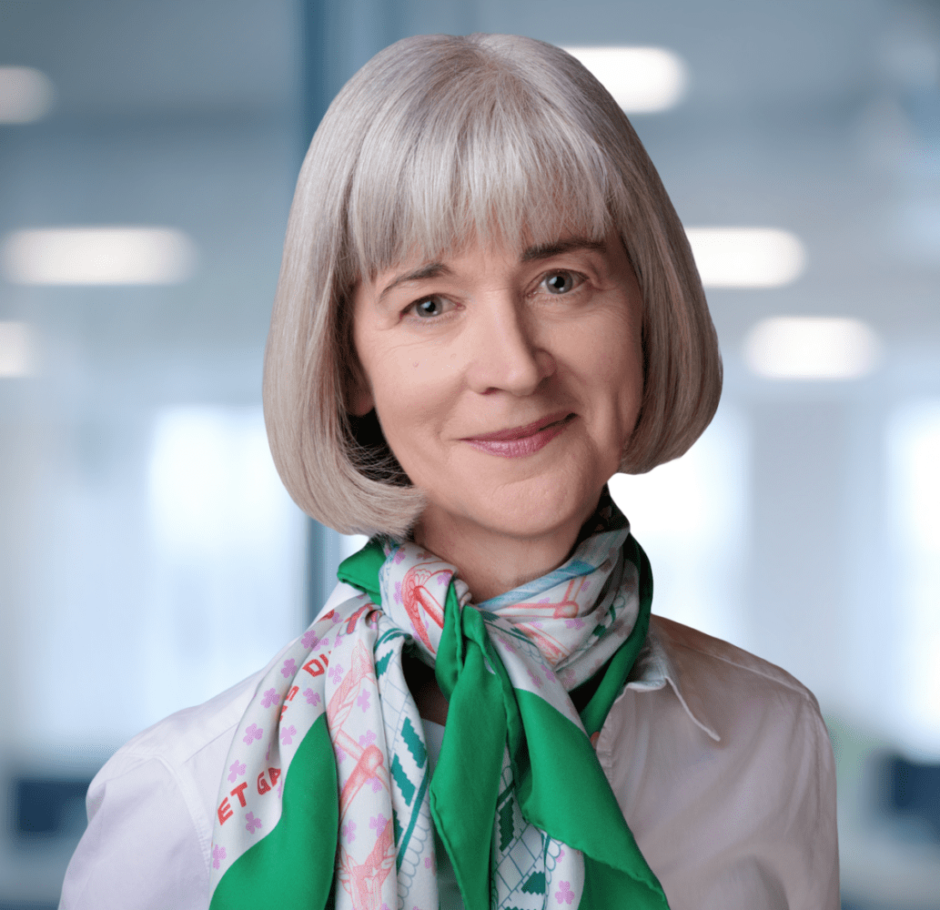 Headshot of a smiling woman with gray hair and a green and pink scarf in a blurred office.