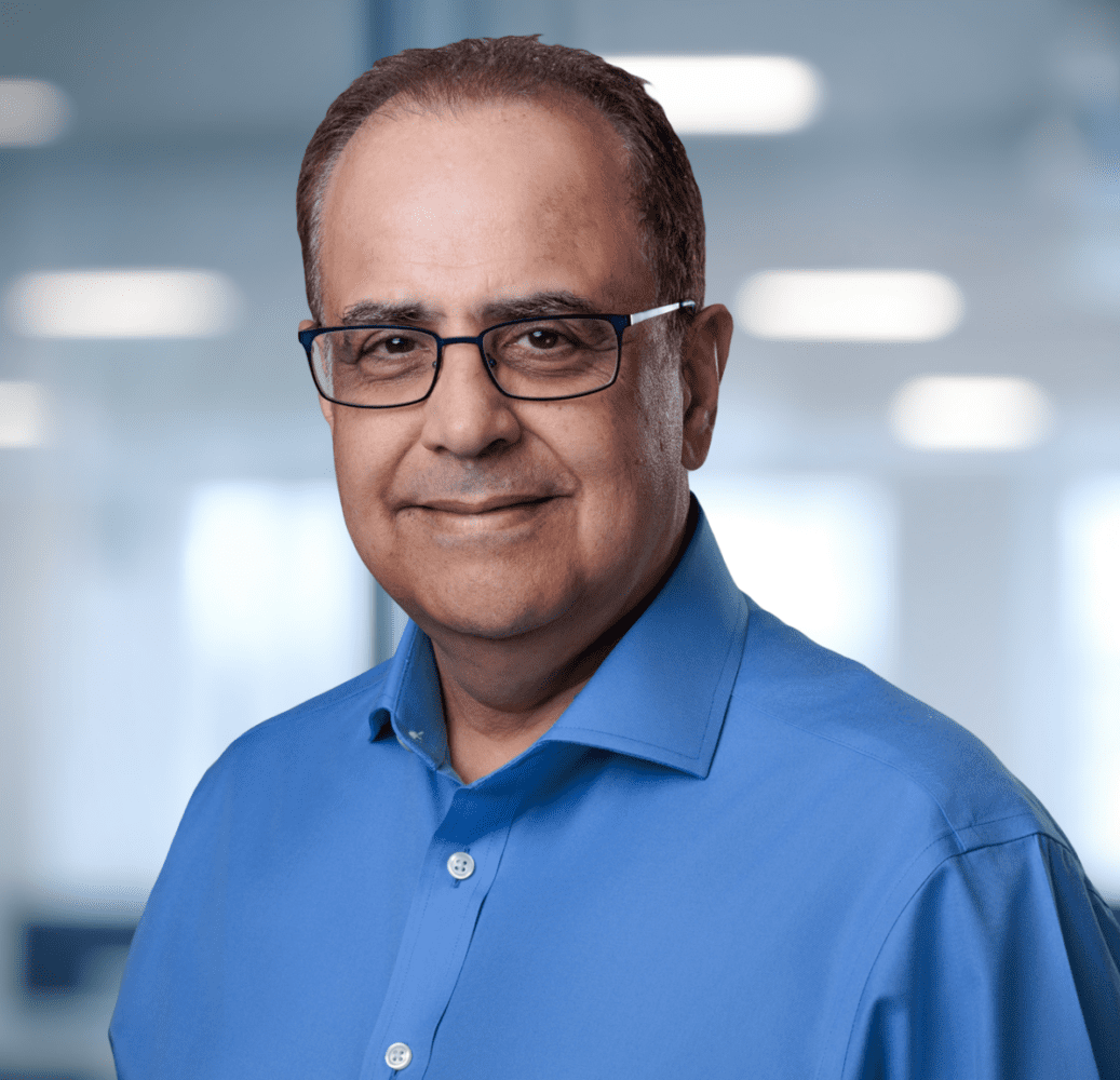 Headshot of a smiling man with glasses and a blue shirt against a blurred office background.