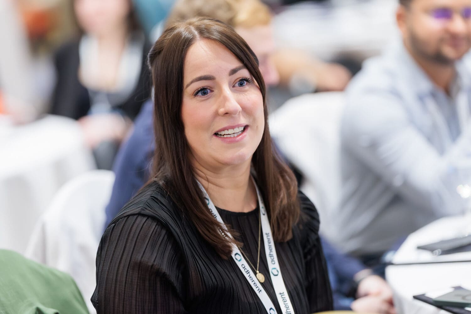 A smiling woman with long brown hair and blue eyes, wearing a black pleated top and an 'adherent' lanyard, looking engaged at a conference.