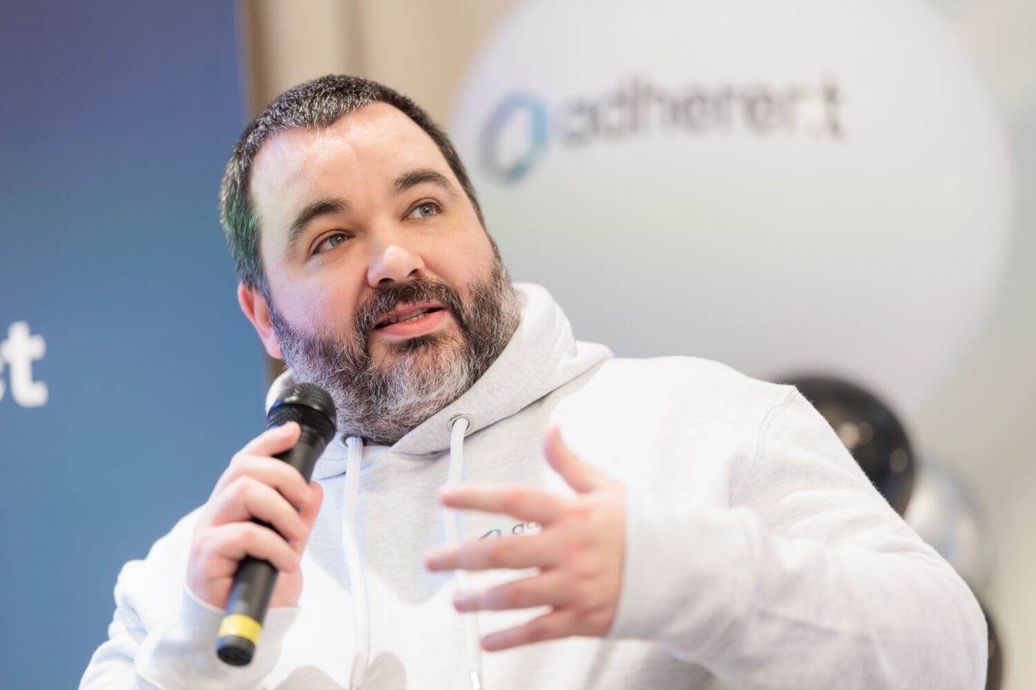A man with a salt-and-pepper beard and light blue eyes speaks into a microphone, gesturing with his right hand. He wears a grey hoodie, and a blurred 'adherent' logo is in the background.