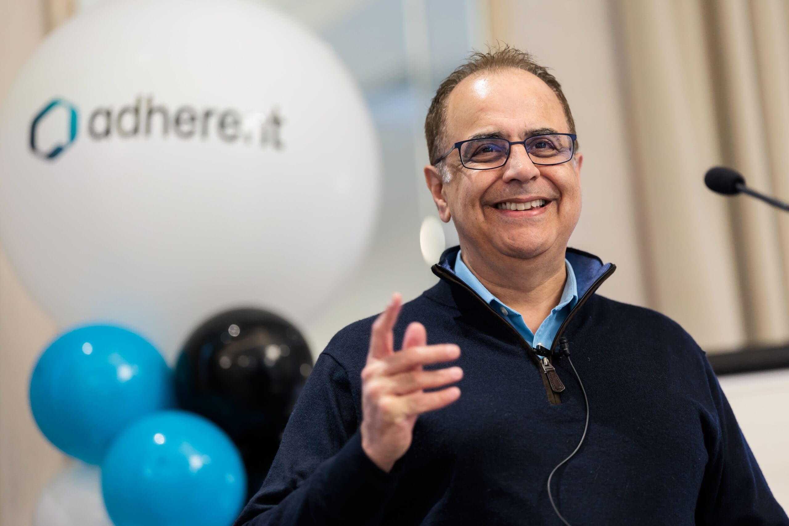A smiling man with glasses and a blue quarter-zip sweater gestures while speaking at an event. A blurred white balloon with 'adhere.it' and other blue and black balloons are in the background.
