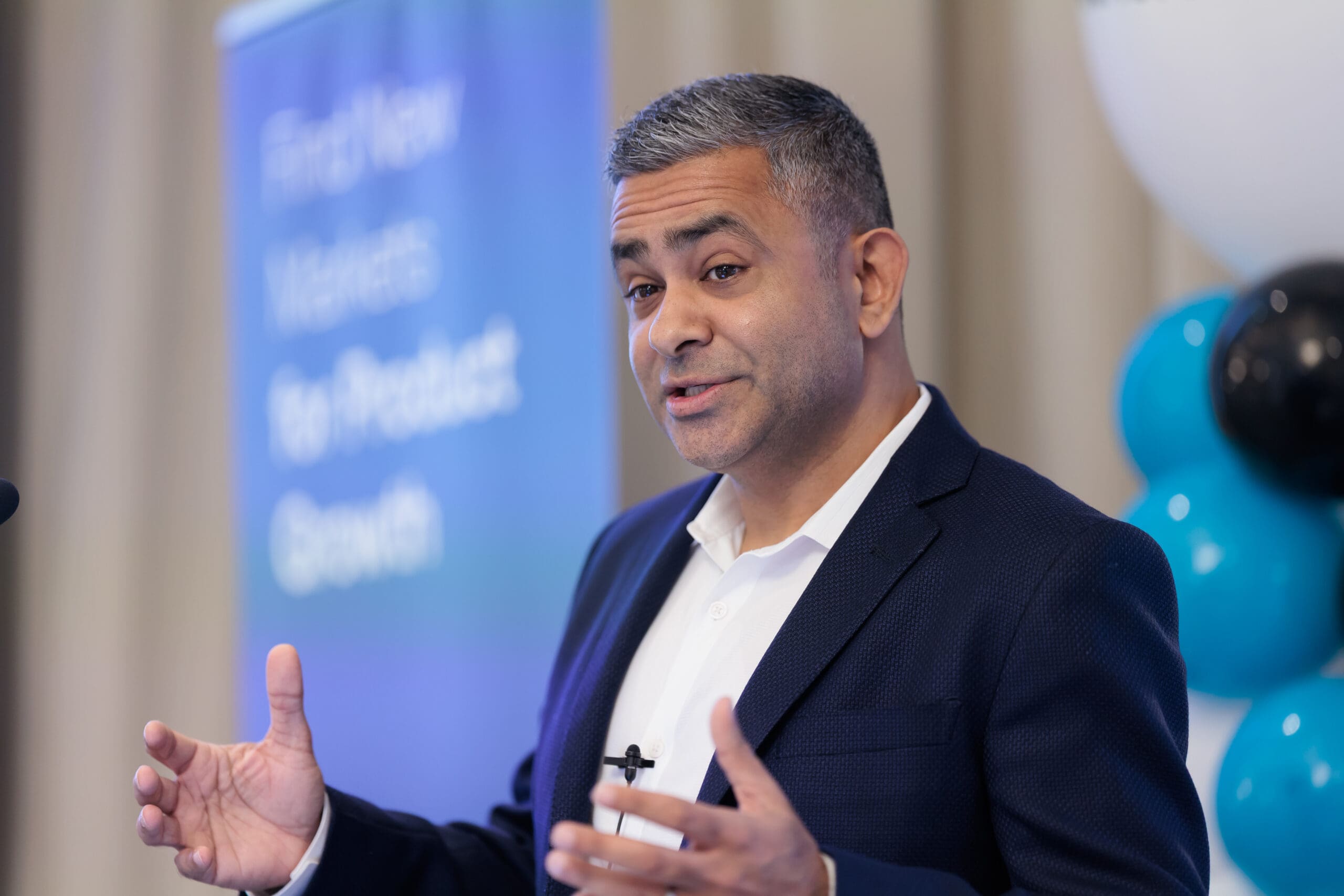 A man with grey-streaked dark hair, wearing a blue suit jacket and white shirt, speaks and gestures with his hand. A blurred blue banner with text 'Find New Markets For Product Growth' is behind him, along with blurred black and blue balloons.