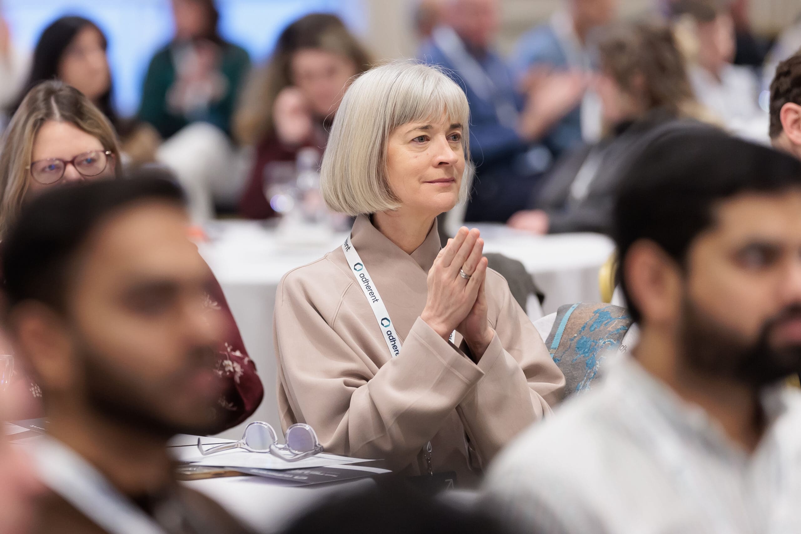 A woman with short gray hair, wearing a beige top and an 'adherent' lanyard, sits with hands clasped, looking thoughtfully to the side at a conference. Blurred attendees are in the foreground and background.