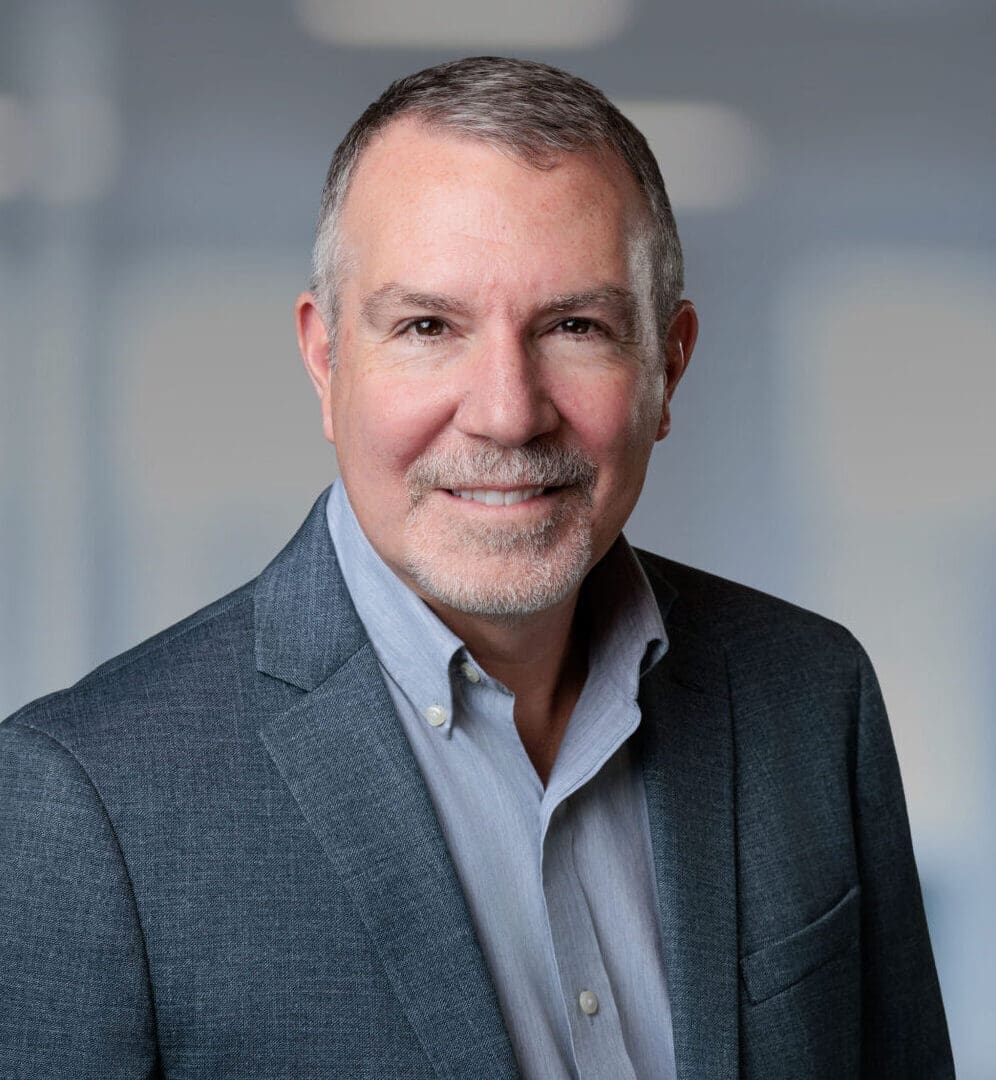 Headshot of a smiling man with grey hair and beard, wearing a grey blazer and blue shirt, in a blurred office background with a green plant.