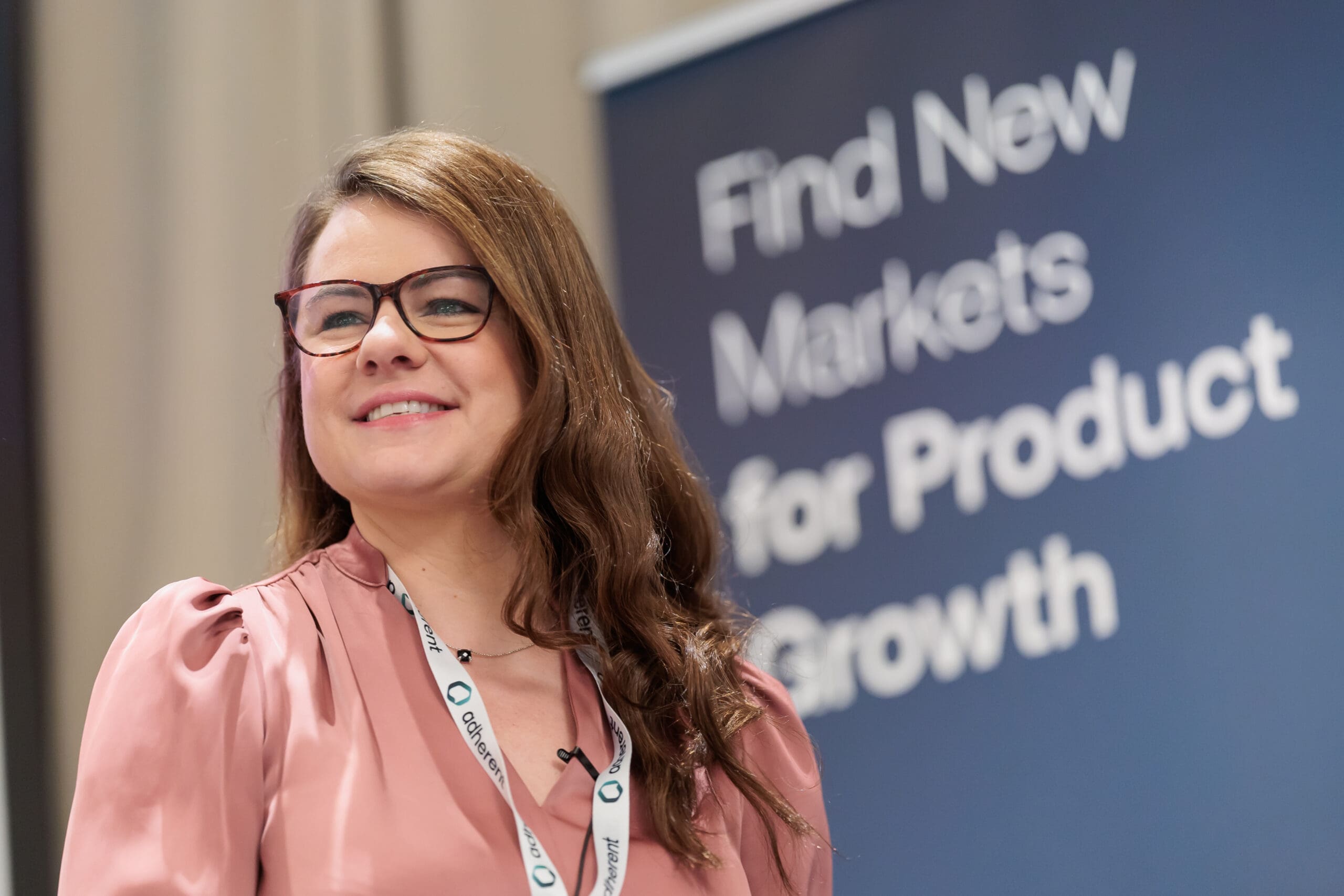 A smiling woman with long brown hair and tortoiseshell glasses, wearing a pink blouse and an "adherent" lanyard. A blurred blue banner behind her reads "Find New Markets for Product Growth."