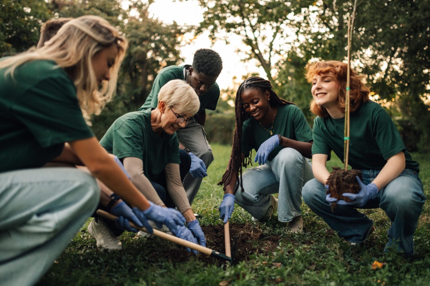 Five diverse volunteers in green shirts and blue gloves smiling and working together to plant a young tree in a grassy outdoor setting.