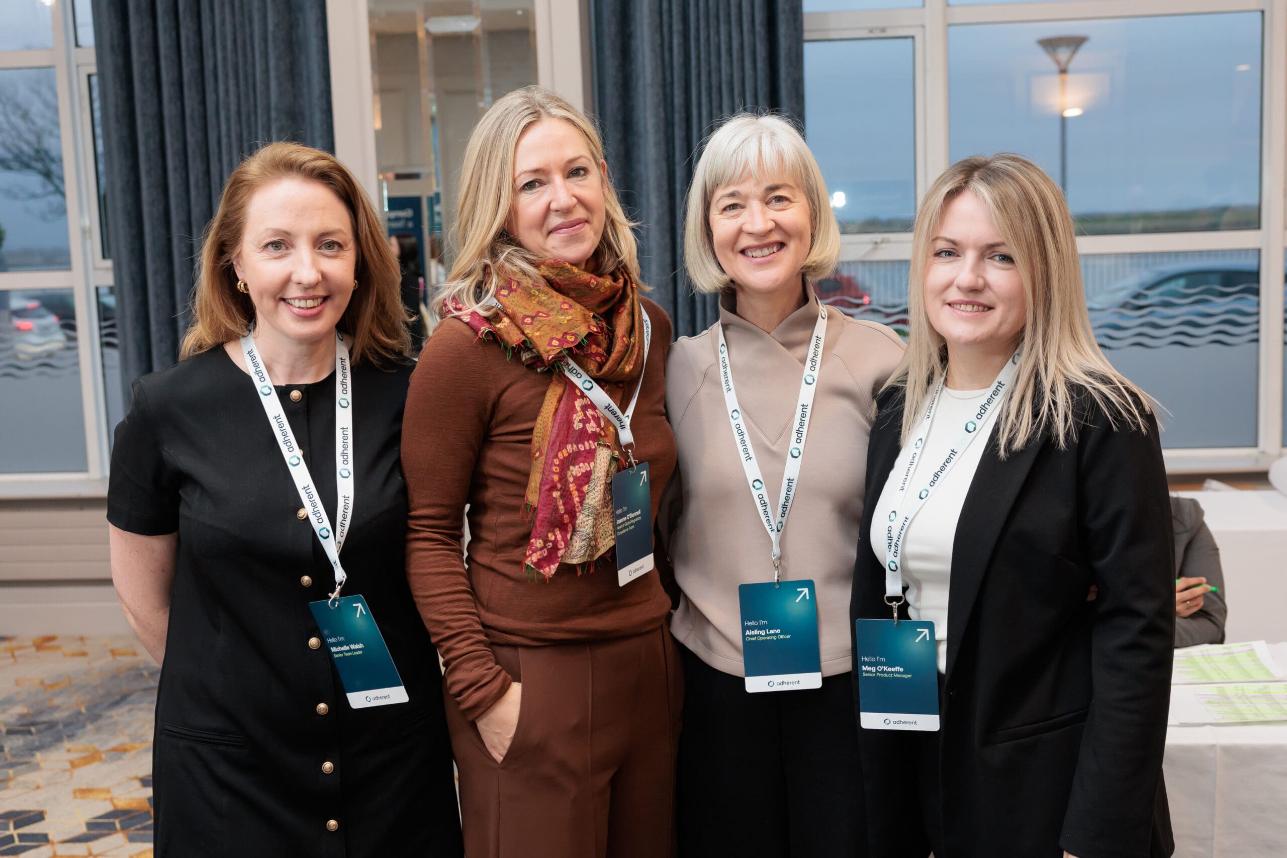Four smiling women from Adherent, wearing lanyards with their names and titles, standing in front of a window at an event.