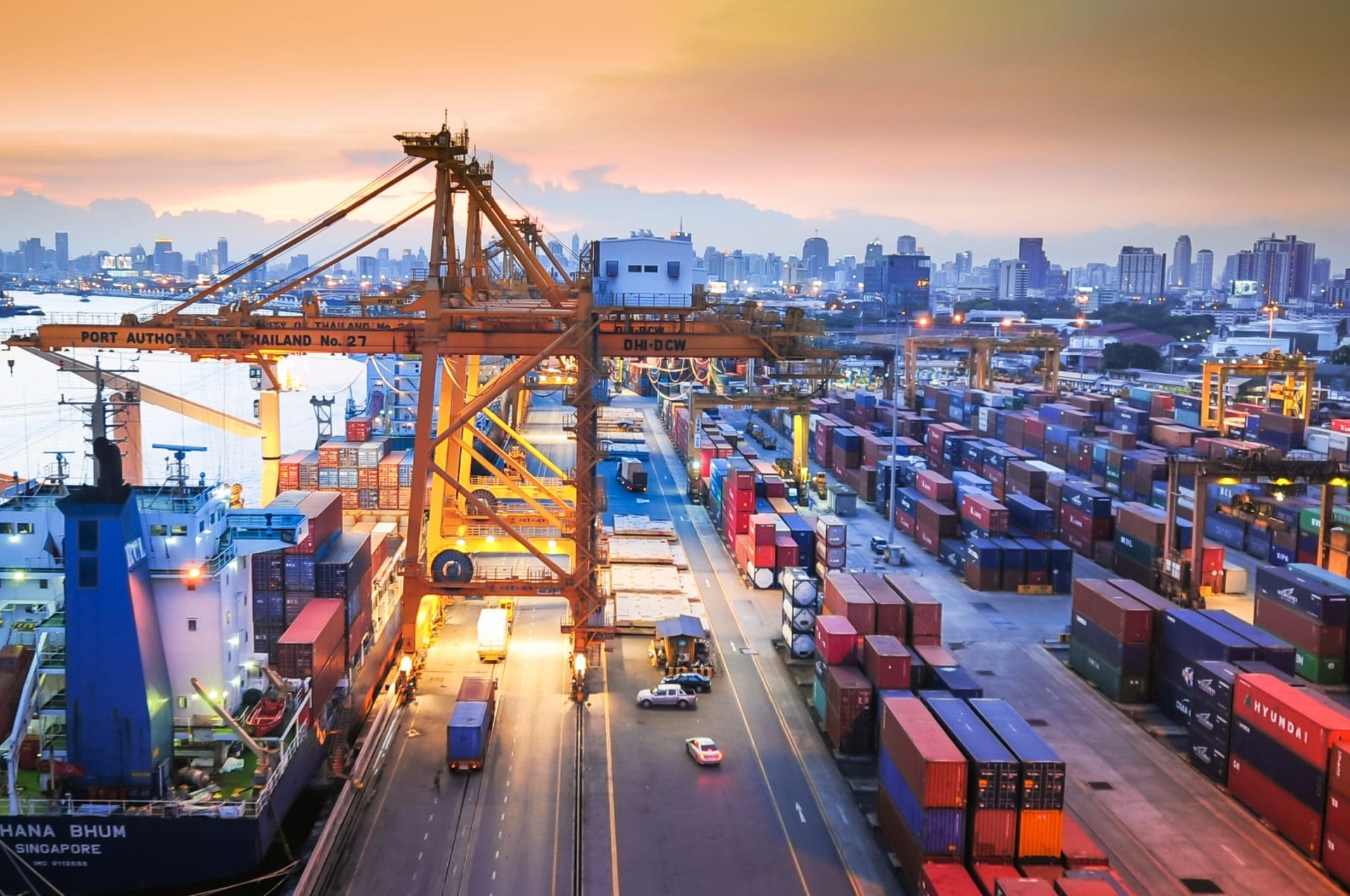 Aerial view of a busy port at dusk, with a container ship, large yellow gantry cranes, rows of stacked shipping containers, and a city skyline in the background under an orange and purple sky.