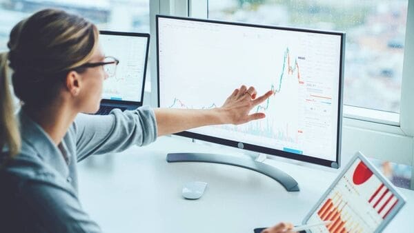 A woman in glasses points at a line graph on a computer monitor, with a laptop and tablet also displaying charts on her desk.