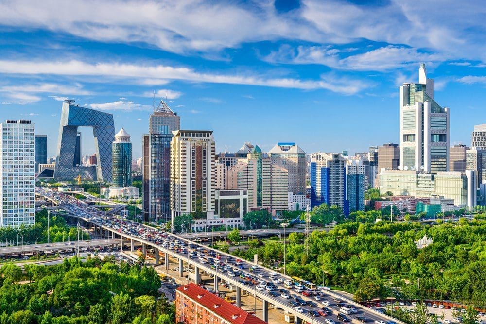 An aerial view of a modern city with tall buildings, including the distinctive CCTV Headquarters, an elevated highway with heavy traffic, and green trees under a blue sky.