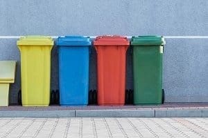 Four colorful recycling bins (yellow, blue, red, green) lined up against a gray wall.