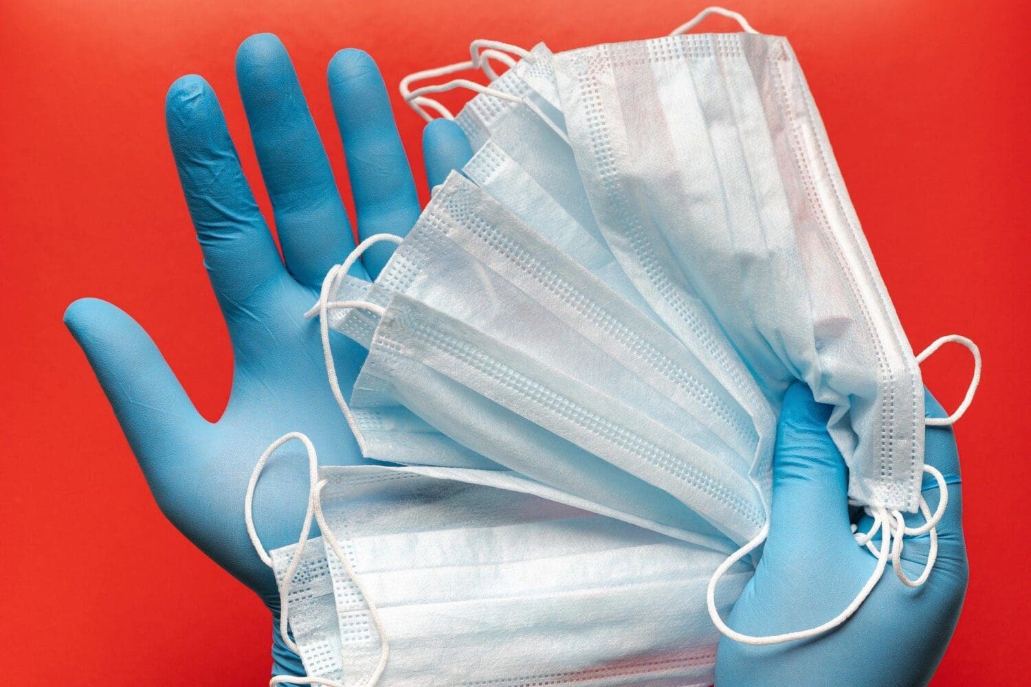 A person wearing blue medical gloves holds a stack of light blue disposable masks against a red background.