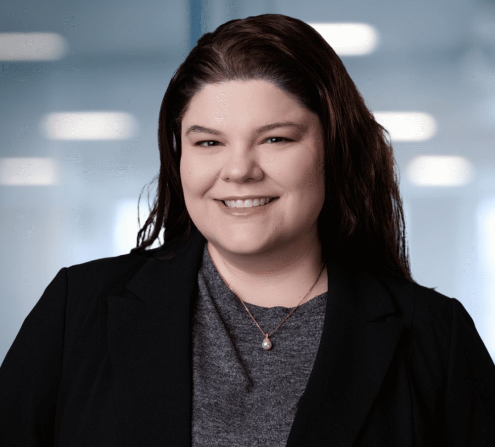 Headshot of a smiling woman with dark brown hair, wearing a black blazer and a pearl necklace, against a blurred office background.
