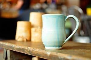 Light blue ceramic mug with a handle on a dark wooden table, with blurred beige objects in the background.