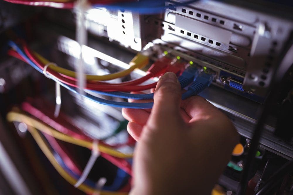 A hand connects blue Ethernet cables to a server rack with multiple colorful cables and illuminated indicator lights.