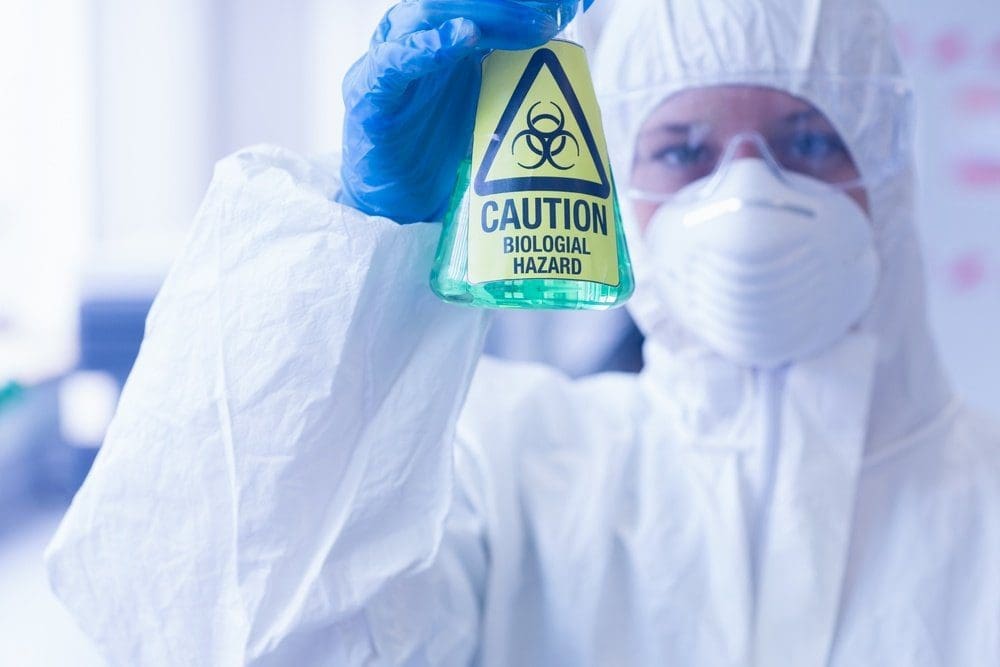 Person in white hazmat suit, blue gloves, and respirator holding a flask with a 'Biological Hazard' label.