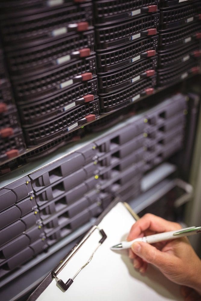 A hand writes on a clipboard in front of rows of dark server racks with illuminated indicator lights.