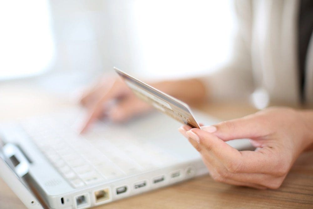 Close-up of a hand holding a gold credit card next to a white laptop keyboard, with another hand blurred on the keyboard.
