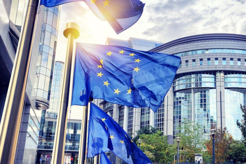 Several European Union flags, blue with yellow stars, wave in front of modern glass buildings under a bright, cloudy sky.
