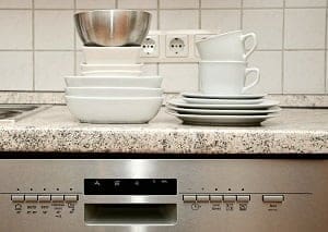 Clean white and silver dishes stacked on a speckled kitchen counter above a stainless steel dishwasher.