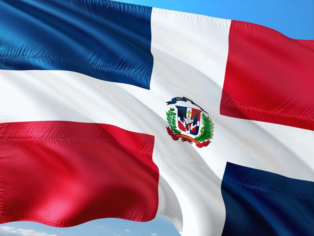 The national flag of the Dominican Republic, with its white cross, blue and red quadrants, and central coat of arms, waving against a blue sky.