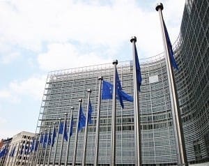 European Union flags on flagpoles in front of a large glass building under a cloudy sky.