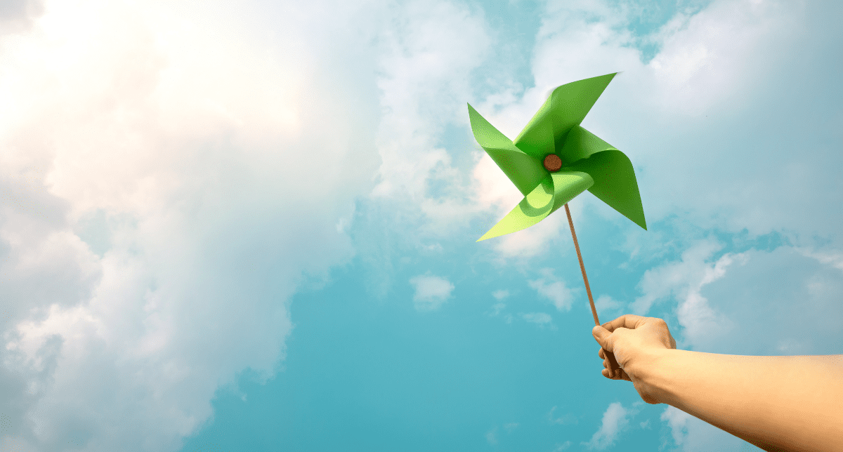 A hand holding a green pinwheel against a bright blue sky with white clouds.