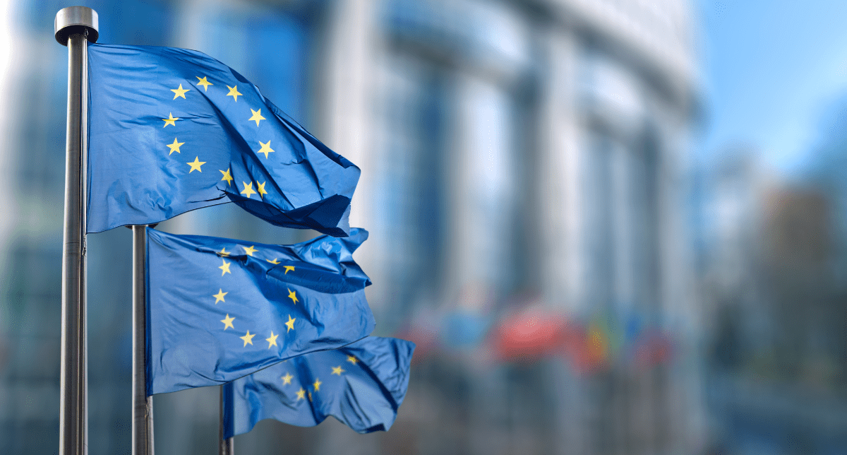 Two blue European Union flags with yellow stars, waving on flagpoles in front of a blurred modern building.