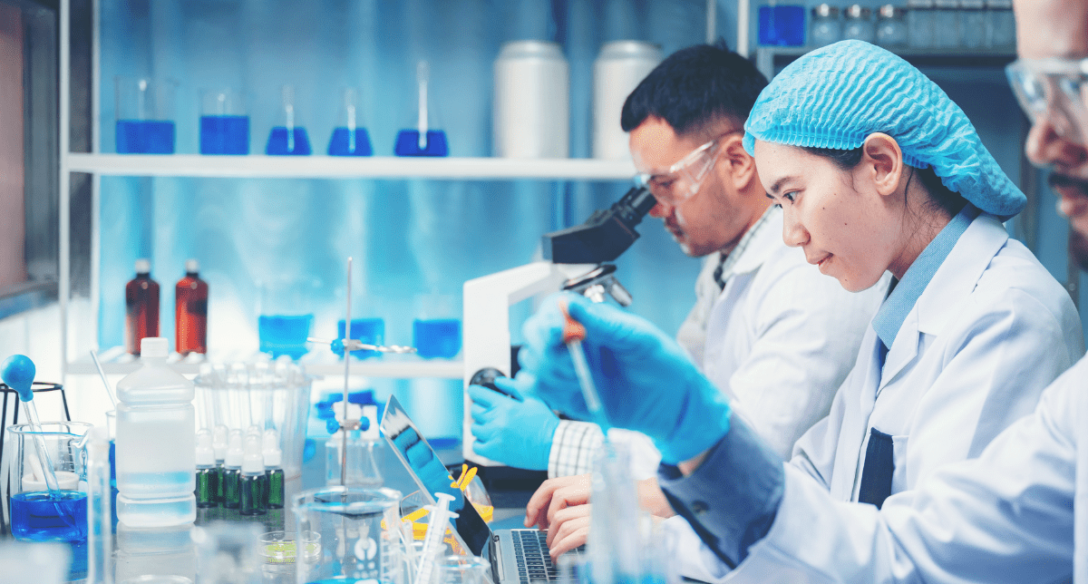 Three scientists in lab coats and safety gear work with microscopes, laptops, and chemical solutions in a laboratory.