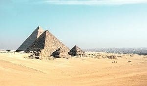 Three ancient pyramids stand in a vast, sandy desert under a clear blue sky, with a distant city visible on the horizon.