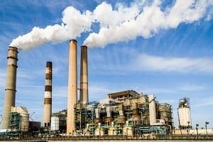 Industrial power plant with multiple smokestacks emitting white smoke against a blue sky.