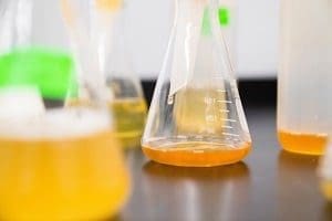 Close-up of laboratory flasks on a dark counter, one containing orange liquid and another with yellow foamy liquid.